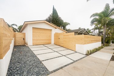 Concrete and gravel walkway leading to a white garage with a wood door surrounded by a wood wall