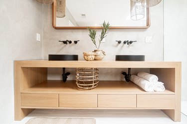 Light wood bathroom vanity with towels, basket, two black sinks, a plant clipping, and a mirror