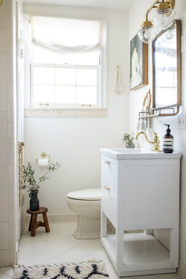 White bathroom with a black-white patterned bathmat, white vanity, wood stool with vase of branches, and multi-sconce