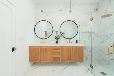 Bathroom with marble walls, gold hardware, pendant lights, round mirrors, wood vanity, and glass door shower