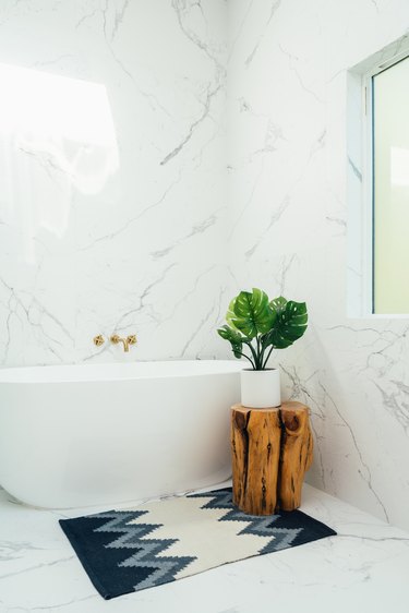 Bathroom with freestanding bathtub, blue-white bath rug, rustic wood stool with a plant and white marble walls