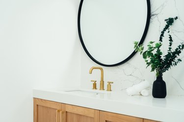 Blond wood bathroom vanity with white counter, gold sink faucet, large round mirror, and black vase with plant clipping