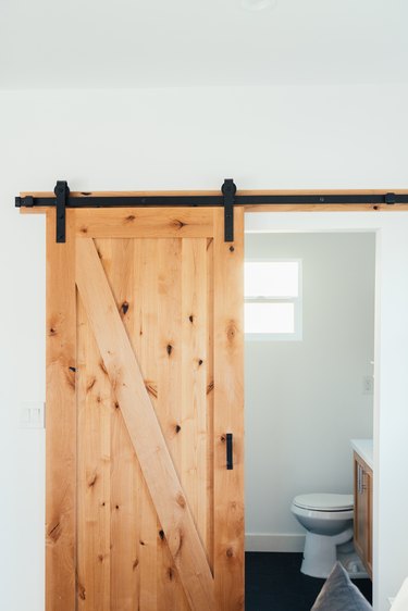 A sliding wood barn door leading to a bathroom with a small square window and toilet.
