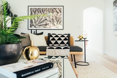 Black living room futon with triangle print throw, loop side table, contemporary art, and a fern plan, gold vase, and books
