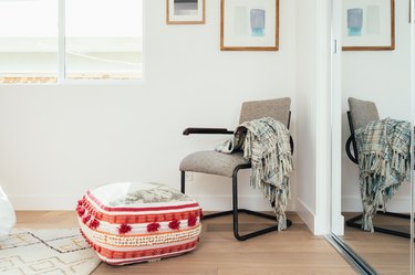 Bedroom corner with light wood floors, fringed boho cushion, and gray armchair with a plaid blanket and abstract art