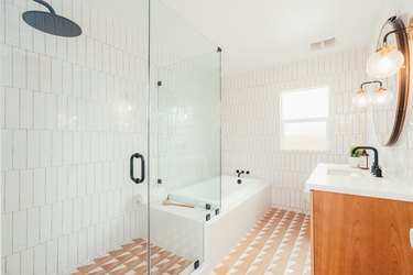 Neutral-white floor tile, white vertical wall tile, alcove tub, glass door shower, light wood vanity, and globe sconces
