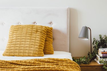 White tufted upholstered headboard with yellow knitted pillows and blankets, wood nightstand with lamp and plant