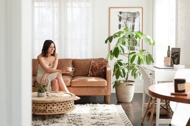 Bright living room with a person sitting on a brown leather couch with an accent pillow. A round stone coffee table, shag rug and plant.