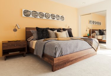 Bedroom with yellow walls, wood night stand, wood headboard, gray-neutral bedding, and mirror closet. An artwork with painted birds.