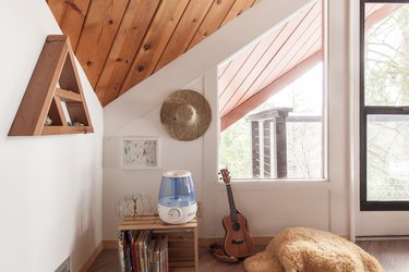 Play room with a triangle shelf, wood crate with books, dehumidifier, hat, guitar. A triangular wood vault ceiling and geometric window.