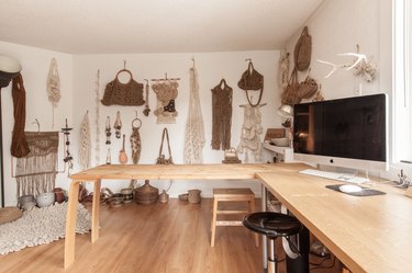 Studio with a display wall of macrame purses and wall hangings. A wood L-shape desk with a computer.