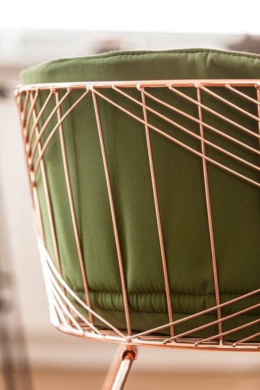 Back of an accent chair with rose gold steel rods, forming a pattern and a green cushion.