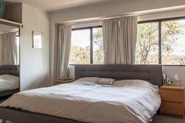 Minimalist bedroom with white bedding and a gray tufted headboard. White dresser night stands are on each side. Gray curtained windows.