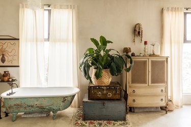 Bathroom with a vintage green tub, vintage suitcases with a plant in a wicker vase. An octopus poster, white vintage dresser, and a mask.
