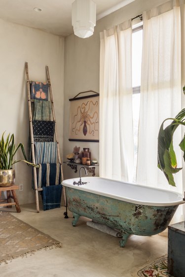 Boho bathroom with rustic clawfoot tub, ladder with towels, art, curtains.