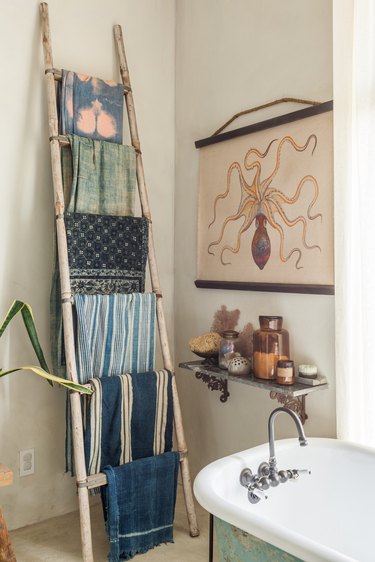Bathroom with a freestanding bathtub. A shelf with bottles, and an octopus poster. A ladder towel rack with eclectic fabrics.