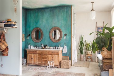 Bathroom with double round mirrors on a turquoise accent wall. Wood vanity with a double sink. Plants and an art deco pendant light.
