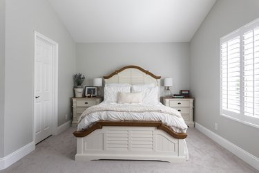 A bed with an ornate wood headboard. White night tables, with clear vase lamps. Light gray walls, with white millwork.