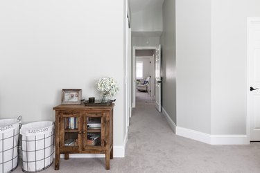 Laundry baskets, next to a wood cabinet. A framed photo and a vase of flowers. Light gray walls, white millwork, and a hallway.