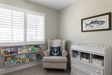 A reading corner with shelves of children's books. A white armchair with an anchor pillow. A framed fish print, over a changing table.