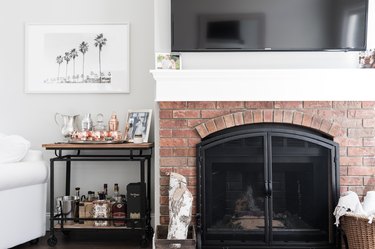 A brick fireplace with baskets on the hearth. A bar cart with copper mule mugs and liquor. A framed photo of palm trees.