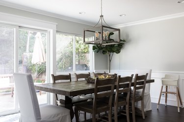 Dining room with dark wood furniture and a box chandelier. The walls are gray, with white wainscoting and millwork. A white highchair.