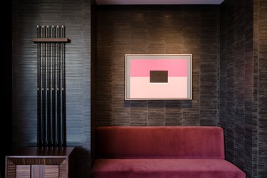 A red sofa with an abstract color block artwork on a dark tile wall. Beside is a rod sculpture and dark wood cubed side table.