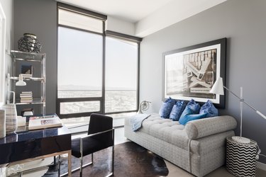 Bright room with gray tufted sofa, blue pillows, zig-zag line coffee side table ,lamp, black chair-desk set, metal shelf, books and sculpture.