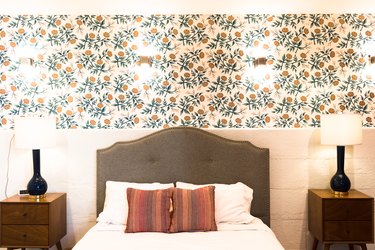 Floral wallpaper and sconces over a bed with a tufted headboard, and colorful accent pillows, two wood nightstands and black table lamps.