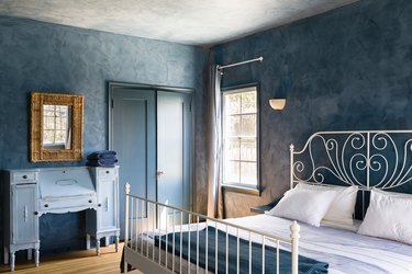 Bedroom with blue walls, white spiral bed frame, blue bedding, vintage blue desk, vintage gold mirror, bifold door, and curtained window.