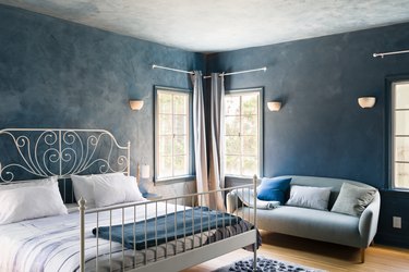 Blue wall bedroom with white spiral bed frame, wall sconces, blue couch, pillows, windows, and wood floor.