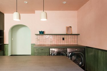 Kitchen with pink walls, pink square tile backslash, green cabinets and shelving, washer/dryer, bell pendant lights, archway.