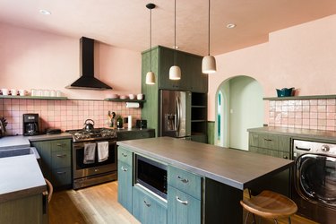 Kitchen with pink walls, green cabinets and shelves, gray counters, pink backsplash, and bell pendant lights.