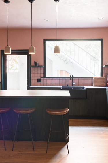 Kitchen with large picture window, pink walls, pink square tile backsplash, green cabinets, wood stools, and bell pendant lights.