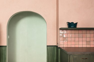 Pink kitchen wall with a pink square tile backsplash, green cabinets, and a green shelf. An arched doorway leading to a light green wall.
