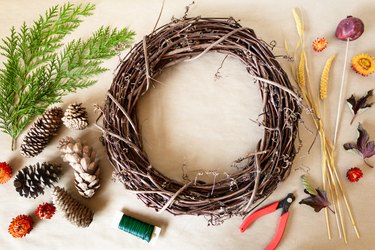 millet stems, cedar branches, wire, wire cutters, dried flowers, pine cones, and a dried pomegranate surround a grapevine wreath base