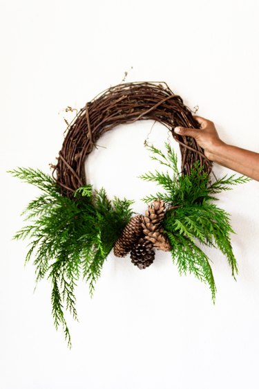 cedar branches and pine cones are attached to a grapevine wreath base