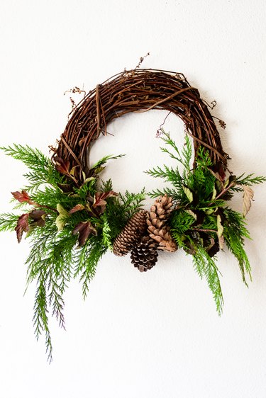 cedar branches and pine cones are attached to a grapevine wreath base