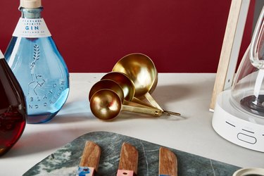 Gold measuring spoons with bottle of gin, marble cutting board, knives, and coffee maker