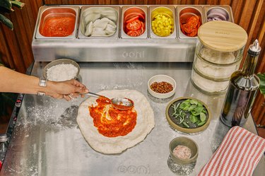 A person spreading tomato sauce on top of pizza dough; on a countertop with containers of toppings