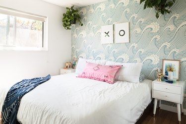 Bedroom with sea wave wallpaper, hanging plants, white nightstand, brass hand sculpture, and Shibori blanket.