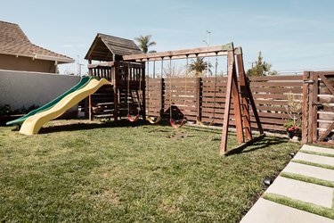 A wooden swing set on top of a patch of grass; a wooden horizontal fence and square concrete pavers