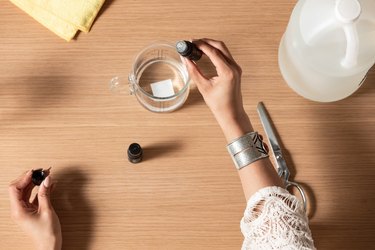 Hands dispensing clear liquid with eyedropper into glass measuring cup next to silver scissors and yellow dryer sheets on wood countertop