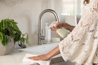 Hand spraying multipurpose cleaner from spray bottle onto sink with silver faucet against marble backsplash with potted plant.