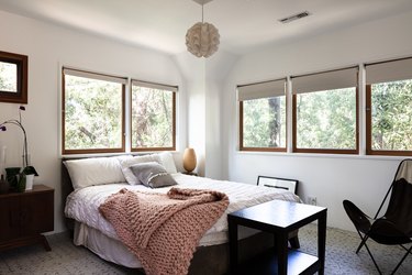 Bedroom with a wave ball pendant light, wood frame windows, white bedding, and a pink crochet blanket.