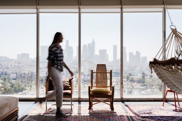 Person in motion, in a living room with a wood rocking chair, colorful traditional rugs, and a hammock. Large windows with a city view.
