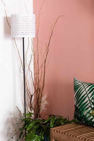Lamp and plant against pink and white-painted wall with slatted wood bench and grass-patterned pillow