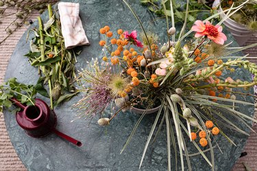 Aerial view of bouquet of pink, purple, and orange flowers with green leaves on round marble table with maroon watering can against tan carpeting