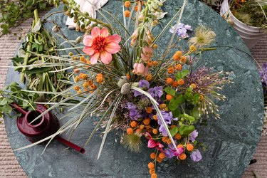 Aerial view of bouquet of pink, purple, and orange flowers with green leaves on round marble table with maroon watering can against tan carpeting