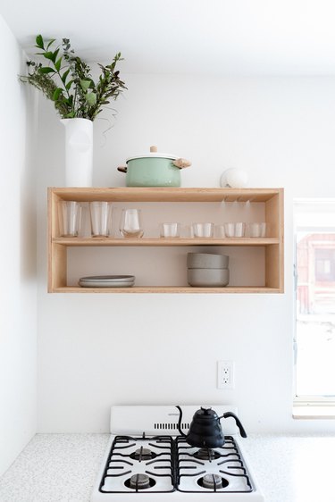 A minimalist white walled kitchen and wood shelving with dishware and plant and apartment size stove